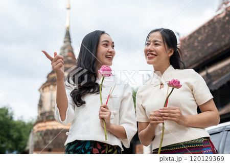Two beautiful, smiling Asian women stand in a temple, enjoying a conversation together. 120129369