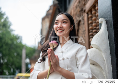 An Asian woman in a traditional dress stands in a temple, holding a lotus in a prayer posture. An Asian woman in a traditional dress stands in a temple, holding a lotus in a prayer posture. 120129395