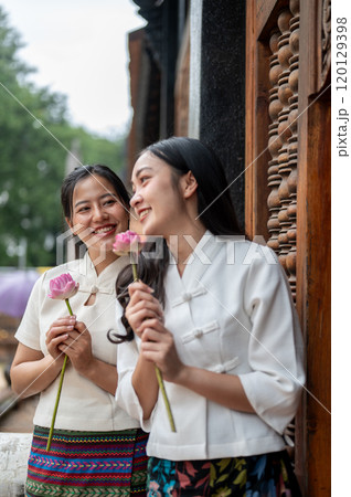 Two charming Asian women in traditional dresses stand in a temple with lotuses in their hand. 120129398