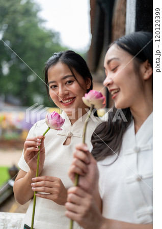 Two charming Asian women in traditional dresses stand in a temple with lotuses in their hand. Two charming Asian women in traditional dresses stand in a temple with lotuses in their hand. 120129399