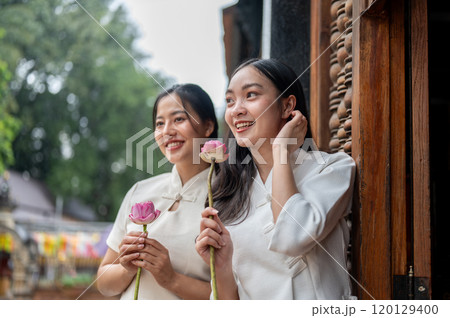 Two charming Asian women in traditional dresses stand in a temple with lotuses in their hand. Two charming Asian women in traditional dresses stand in a temple with lotuses in their hand. 120129400