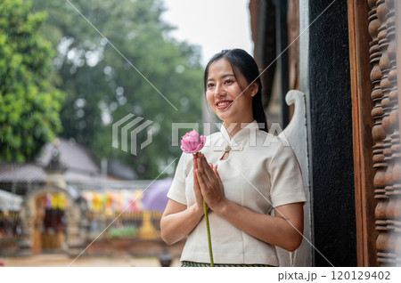 An Asian woman in a traditional dress stands in a temple, holding a lotus in a prayer posture. 120129402