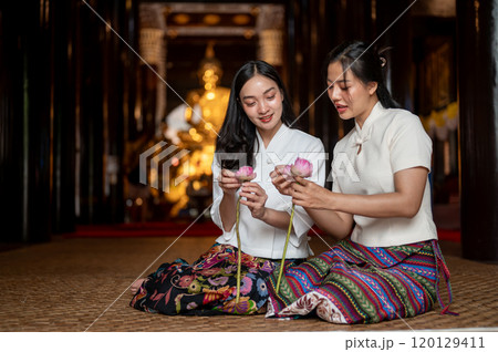 Two beautiful Asian women in traditional dresses sit in a temple, enjoy folding lotus flower petals. Two beautiful Asian women in traditional dresses sit in a temple, enjoy folding lotus flower petals. 120129411
