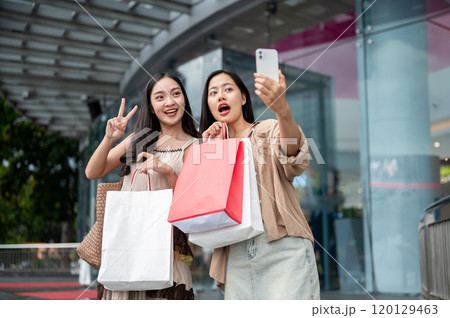 Cheerful Asian female friends enjoy taking selfies together after their shopping spree at the mall. 120129463