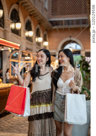 Two charming Asian women enjoying shopping together at a mall, carrying their shopping bags. 120129506