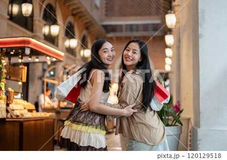 Two attractive Asian female friends enjoy shopping together at a mall, smiling at the camera. 120129518