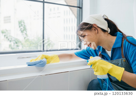 A maid with protective gloves wipes ceramic tiles in a bathroom using a microfiber cloth. Her routine cleaning highlights the essence of purity and hygiene in commercial cleaning. 120133630
