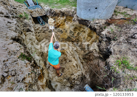 While manually digging pit for septic tank, worker throws soil into garden wheelbarrow using hand shovel. 120133953