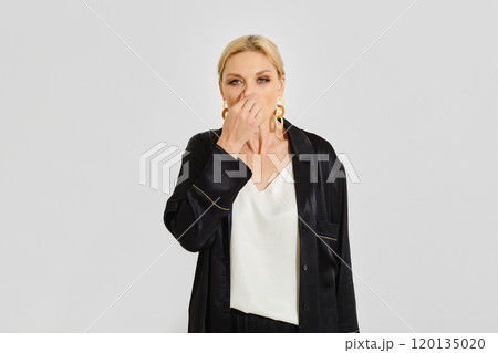 Woman with a cold wipes her nose with her hand standing against a neutral backdrop 120135020