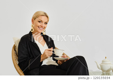A woman enjoys tea in a stylish outfit while seated in a cozy corner with elegant decor 120135034