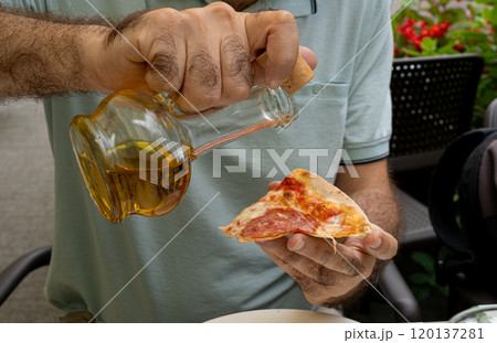 Hand pours olive oil from a bottle onto a slice of pizza at outdoors cafe, summer greenery, casual seating 120137281