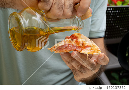 Hand pours olive oil from a bottle onto a slice of pizza at outdoors cafe, summer greenery, casual seating 120137282