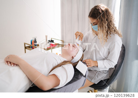 The beautician wearing a protective mask performs a detailed skincare treatment on a client, emphasizing cleanliness and care. The beautician wearing a protective mask performs a detailed skincare treatment on a client, emphasizing cleanliness and care. 120139628