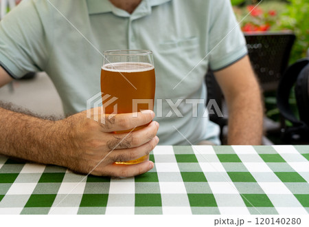 Man holding a glass of beer, sitting at a table with a green checkered tablecloth, outdoors at a cafe 120140280