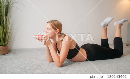 Calorie cake. Sweet craving. Relaxed smiling young lady lying on mat in black sportswear smelling cake in morning at home. Calorie cake. Sweet craving. Relaxed smiling young lady lying on mat in black sportswear smelling cake in morning at home. 120143401