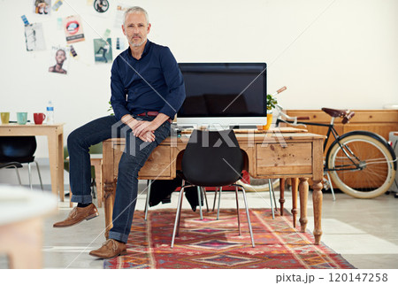 Hes a big player in the business. Portrait of a casually-dressed mature man sitting on his desk in an office. Hes a big player in the business. Portrait of a casually-dressed mature man sitting on his desk in an office. 120147258