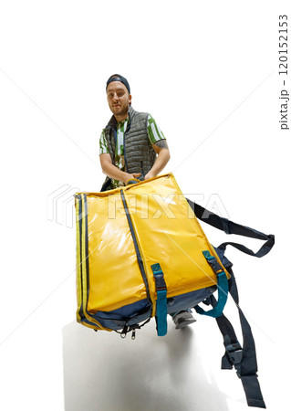 Wide angle photo of young man, courier, dressed casually and carrying huge food delivery pack against white studio background. 120152153