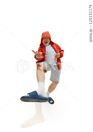 Pensioner, dressed red jacket, shorts and slippers dancing raising legs against white studio background. Excited mature man. 120152174
