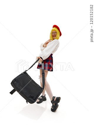 Woman in quirky schoolgirl outfit with yellow wig, red beret and plaid skirt posing playfully with bag against white studio background. Woman in quirky schoolgirl outfit with yellow wig, red beret and plaid skirt posing playfully with bag against white studio background. 120152182