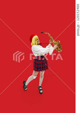 Happy, excited woman dressed stylish vibrant outfit with yellow wig posing with saxophone against vibrant red background. 120152460