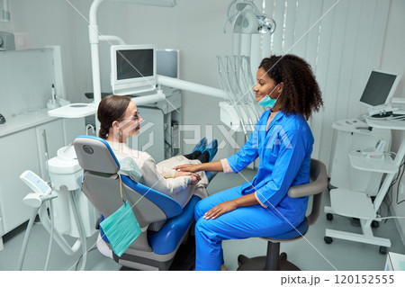Dentist sits beside patient in dental chair, gently holding her hand and offering reassuring smile before procedure. Dentist sits beside patient in dental chair, gently holding her hand and offering reassuring smile before procedure. 120152555