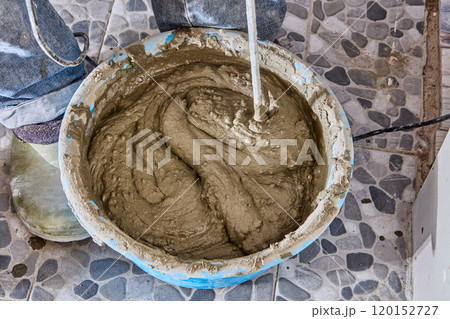 Construction worker tiler mixes cement adhesive mortar in bucket using paddle drill attachment. 120152727