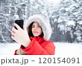 Beautiful brunette in winter in forest in red jacket with hood on her head with smartphone in hand. 120154091