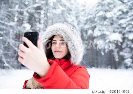 Beautiful brunette in winter in forest in red jacket with hood on her head with smartphone in hand. 120154091