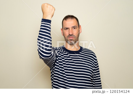 Studio portrait of a happy and overjoyed bearded hispanic man in his 40s, making a winner gesture and raising his fist in celebration, isolated over a beige background. Lifestyle concept. Studio portrait of a happy and overjoyed bearded hispanic man in his 40s, making a winner gesture and raising his fist in celebration, isolated over a beige background. Lifestyle concept. 120154124