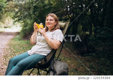 Happy smiling 30s women is holding thermos mug and sitting on deck chair in forest. Hot tea or other beverage on summer day. Lifestyle adventure concept. Woman drink cold water from the bottle. 120154364