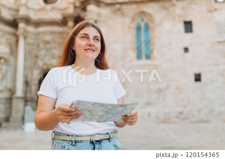 Attractive female tourist is exploring new city. Redhead woman holding a paper map near the Virgin Saint Mary, Valencia Cathedral. Traveling Europe 120154365
