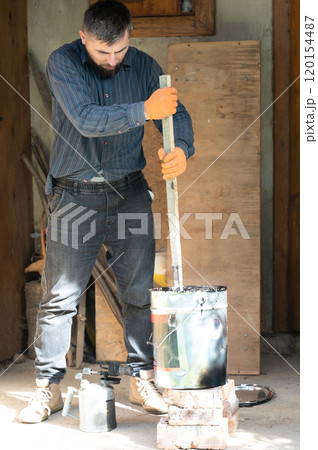 A man stirs liquid bitumen in a bucket with a wooden stick. 120154487