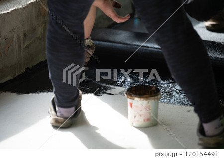 A woman uses a brush to apply bitumen to a concrete floor. A woman uses a brush to apply bitumen to a concrete floor. 120154491