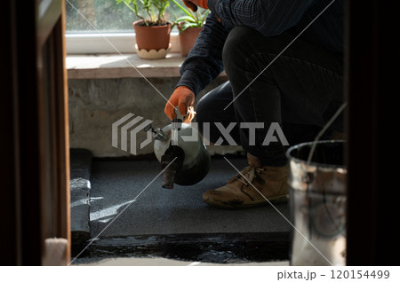 A man uses a kerosene lamp to heat a roll of bitumen for waterproofing the floor. 120154499