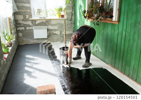 A woman uses a brush to apply bitumen to a concrete floor. A woman uses a brush to apply bitumen to a concrete floor. 120154501