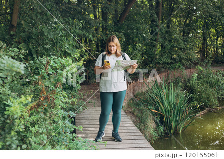 Stylish hipster woman holding paper map and phone, wearing backpack and relaxing in nature. Travel and wanderlust concept. Amazing chill moment, summer time. Full body 120155061