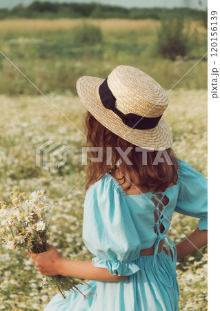 Girl in blue linen dress and yellow knitted kerchief stands back and holds bouquet of flowers in daisy field close up Girl in blue linen dress and yellow knitted kerchief stands back and holds bouquet of flowers in daisy field close up 120156139