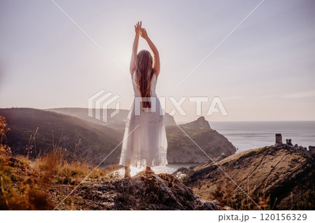 A woman stands on a hill overlooking the ocean. She is wearing a white dress and has her hands on her head. The scene is serene and peaceful, with the woman looking out over the water. A woman stands on a hill overlooking the ocean. She is wearing a white dress and has her hands on her head. The scene is serene and peaceful, with the woman looking out over the water. 120156329