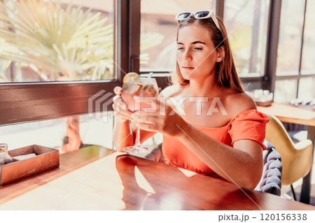 woman in a cafe seated, holding beverage. Dressed in an orange dress with her hair down. Bright interior, large windows allow natural light. Beverage consumption for refreshment. woman in a cafe seated, holding beverage. Dressed in an orange dress with her hair down. Bright interior, large windows allow natural light. Beverage consumption for refreshment. 120156338
