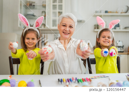 Happy Easter. Two little girls sisters twins grandmother painting eggs enjoying time together. Happy family grandma granddaughters child kids preparing for Easter. Spring Christian festival tradition 120156447