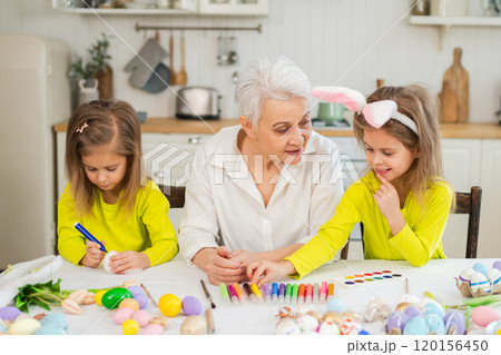 Happy Easter. Two little girls sisters twins grandmother painting eggs enjoying time together. Happy family grandma granddaughters child kids preparing for Easter. Spring Christian festival tradition 120156450
