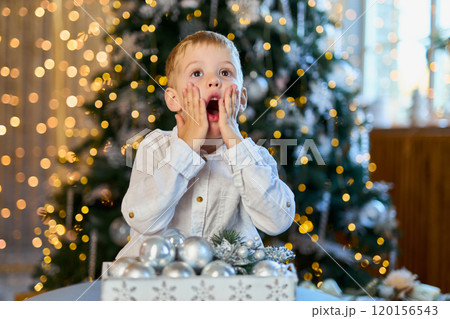 Little child boy decorates Christmas fir tree with silver ornament balls. Small kid holding box with Little child boy decorates Christmas fir tree with silver ornament balls. Small kid holding box with 120156543