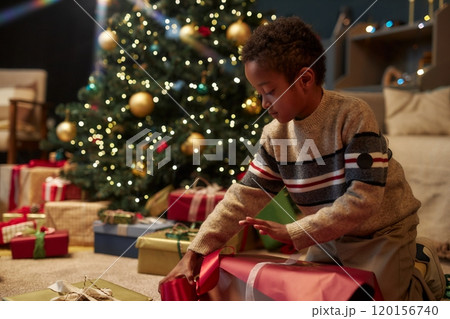 Side view portrait of little African American boy opening Christmas presents and ripping wrapping paper while celebrating holidays and enjoying surprise copy space 120156740