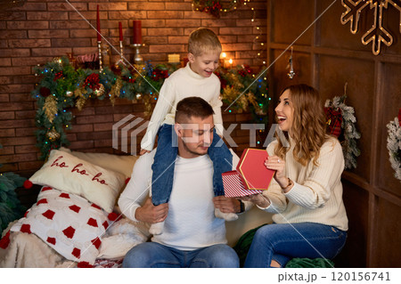 Happy family opening presents next to Christmas tree and smiling cheerfully on Christmas Eve in a 120156741
