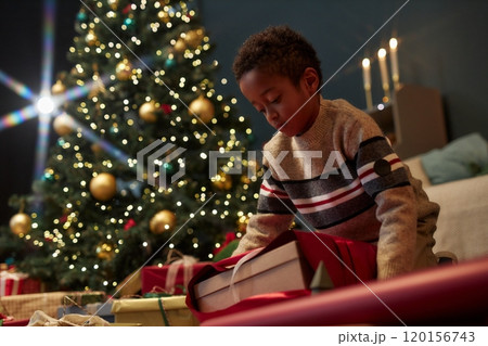Low angle portrait of young African American boy opening Christmas presents in elegant home setting with twinkling lights copy space 120156743