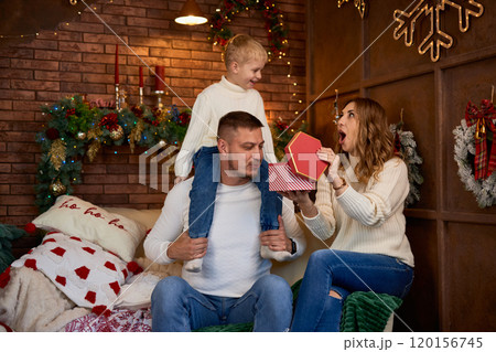 Happy family opening presents next to Christmas tree and smiling cheerfully on Christmas Eve in a Happy family opening presents next to Christmas tree and smiling cheerfully on Christmas Eve in a 120156745