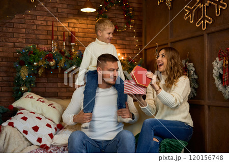 Happy family opening presents next to Christmas tree and smiling cheerfully on Christmas Eve in a 120156748