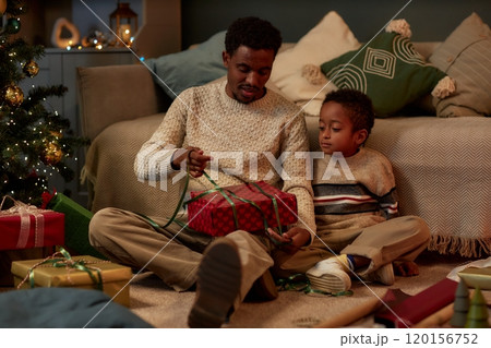 Full length portrait of happy African American father and son opening Christmas presents together sitting on floor in dim lighting and celebrating holidays copy space Full length portrait of happy African American father and son opening Christmas presents together sitting on floor in dim lighting and celebrating holidays copy space 120156752