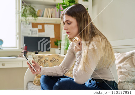 Young serious woman looking at smartphone sitting on couch at home 120157951