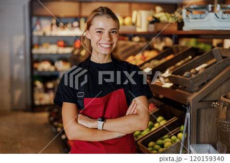 A cheerful female grocery employee stands in the produce section, ready to assist customers 120159340